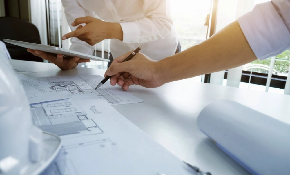 Immagine tematica con tavolo di lavoro in cui progettisti lavorano su disegni tecnici e usando penne e tablet