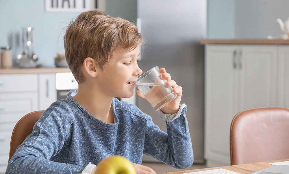 Bambino di profilo che beve un bicchiere d'acqua, sullo sfondo si intravede la cucina di una casa luminosa.